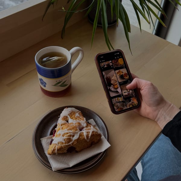 A scone with icing and a cup of tea on a wooden table, with a person holding a phone.
