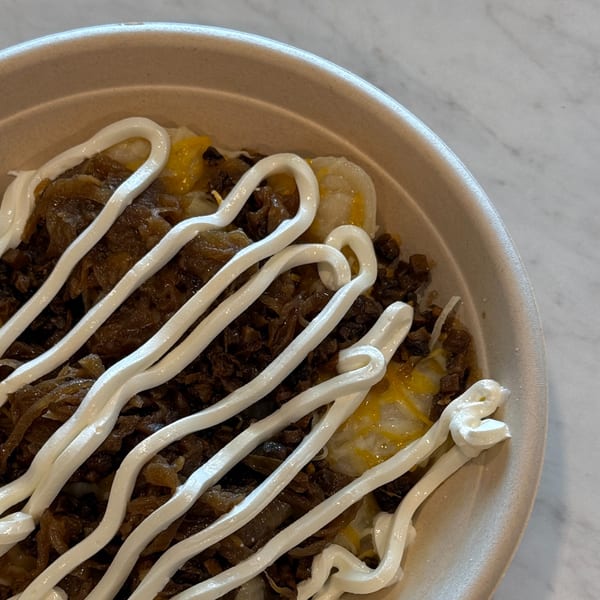 Close-up of a cafe bowl with dark meat, caramelized onions, and white sauce drizzled on top.