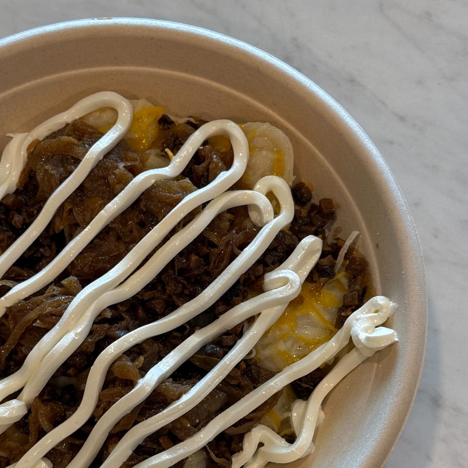 Close-up of a cafe bowl with dark meat, caramelized onions, and white sauce drizzled on top.