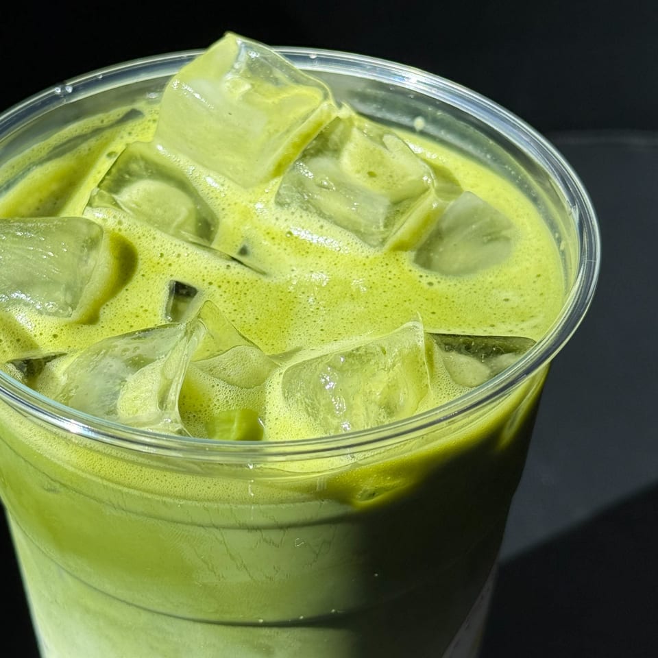 Close-up of an iced matcha latte with foam and ice cubes in a clear plastic cup.