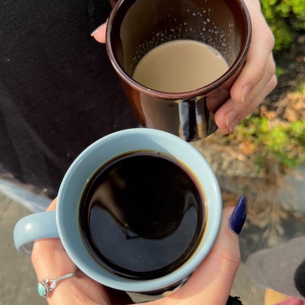 Two hands holding mugs of coffee, one black and one with cream, outdoors at Sunny Cafe.
