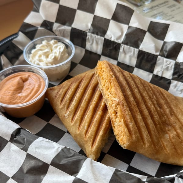 Two golden-brown grilled sandwiches with fillings, served in a basket with checkered paper and two dipping sauces.