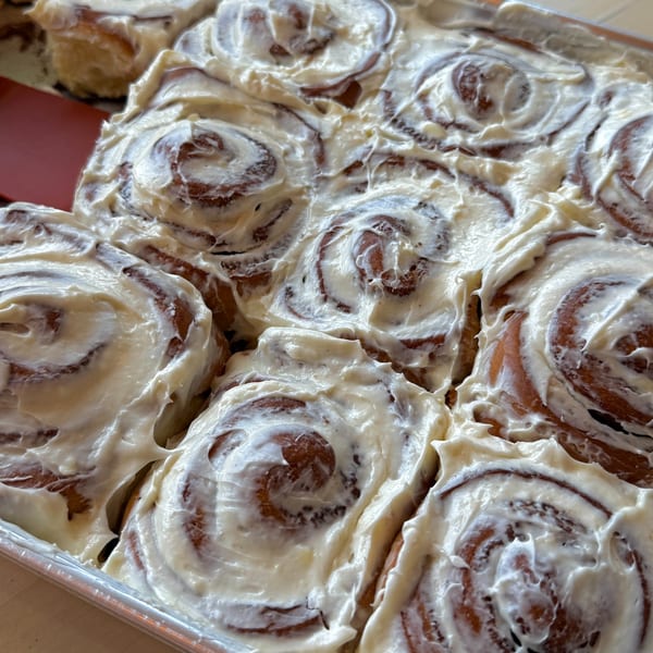 A tray of freshly baked cinnamon rolls, generously covered with white cream cheese frosting.